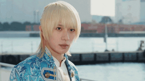 a young man with blonde hair and a blue jacket stands in front of a body of water