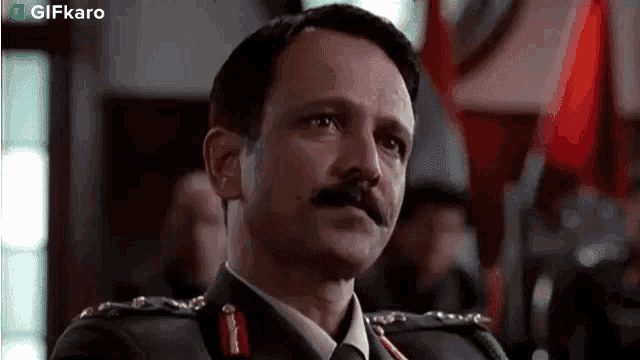 a man in a military uniform with a mustache is sitting in front of a flag .