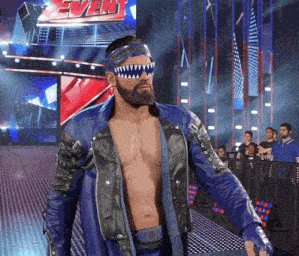 a shirtless wrestler in a blue jacket stands in front of a sign that says even