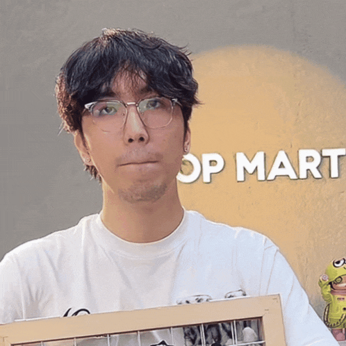 a man wearing glasses and a white shirt holds a picture frame in front of a sign that says op mart