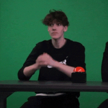 a young man is sitting at a table with a green screen behind him .