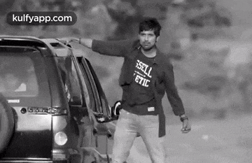 a man wearing a t-shirt that says sell athletic is standing in front of a car .