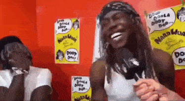a man is laughing while holding a microphone in front of a red wall with posters on it .