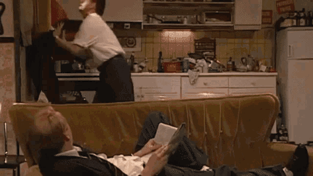 a man sits on a couch reading a book while another man stands in the kitchen