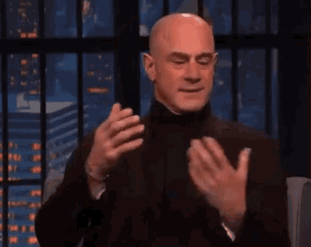 a bald man in a suit is clapping his hands while sitting in front of a fence .