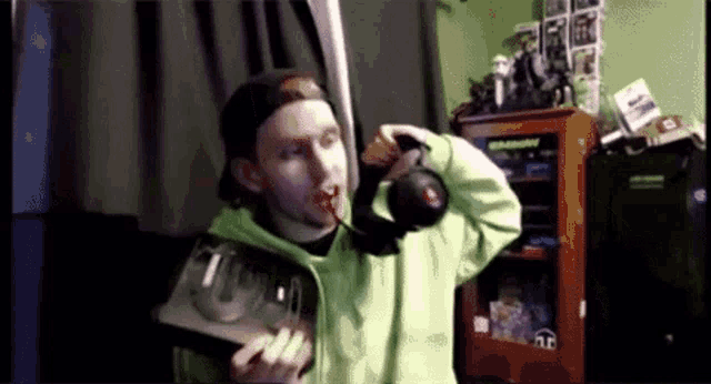 a man in a green hoodie is holding a video game controller and drinking from it .
