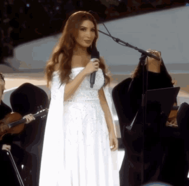 a woman in a white dress is singing into a microphone in front of an orchestra