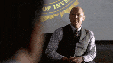 a man in a vest and tie sits in front of a sign that says ' department of investigation '