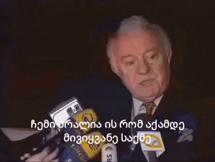 a man in a suit and tie is speaking into a microphone with foreign writing on the screen