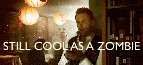 a man is standing in front of a bookshelf holding a cell phone and the words still cool as a zombie are above him .