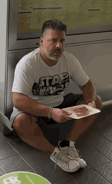 a man wearing a star wars shirt is sitting on the floor holding a tray of food