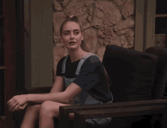 a woman in overalls sits in a chair with a stone wall behind her