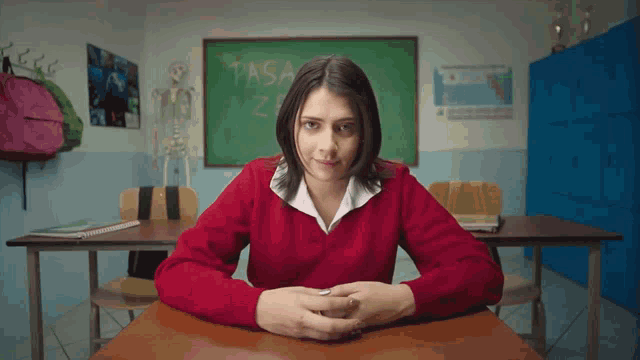 a girl in a red sweater sits at a desk in front of a blackboard that says tasa