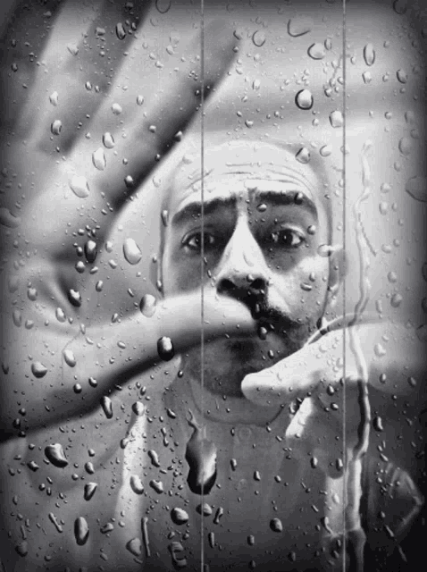 a black and white photo of a man behind a window with rain drops