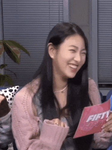 a woman in a pink sweater is sitting on a couch holding a pink card .