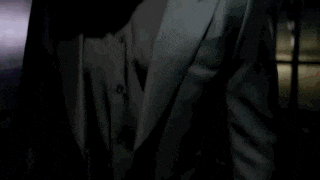 a man in a suit and tie is standing in a dark room with a shadow on his face .