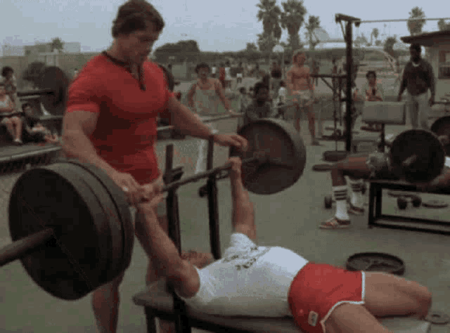 arnold schwarzenegger is helping a man lift a barbell while laying on a bench .