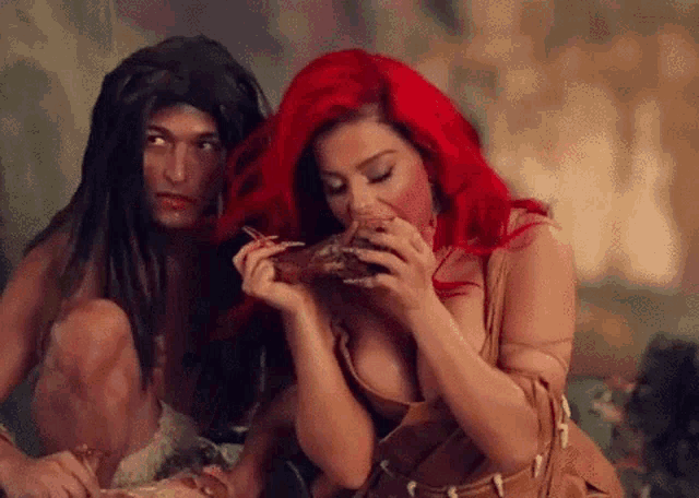 a woman with red hair is eating a piece of meat next to a man with long black hair .