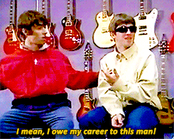 two men are sitting in front of a wall of guitars and one says " i mean i owe my career to this man ! "