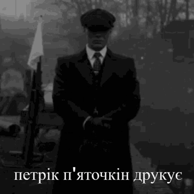 a black and white photo of a man in a suit and hat standing in front of a flag .