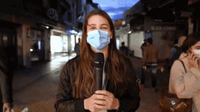 a woman wearing a mask is holding a microphone in front of a black corner store