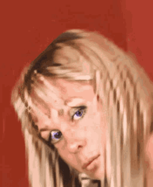 a woman with blonde hair and blue eyes looks at the camera with a red background