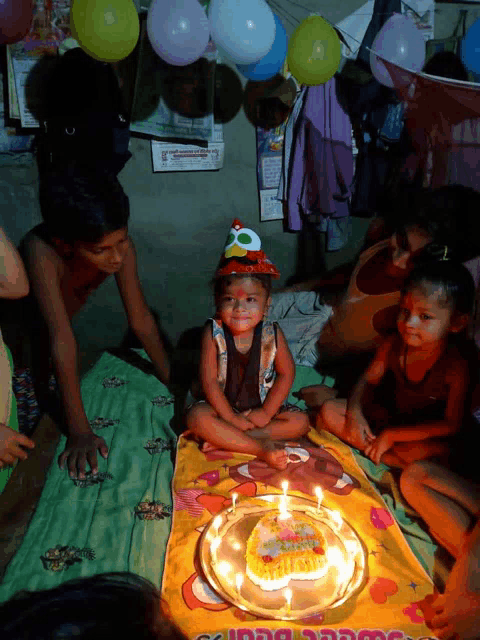 a little boy wearing a birthday hat sits on a blanket next to a cake with candles