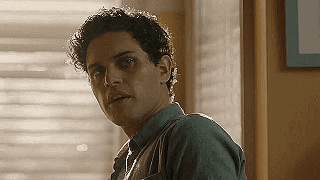 a man with curly hair is standing in front of a window in a room and looking at the camera .