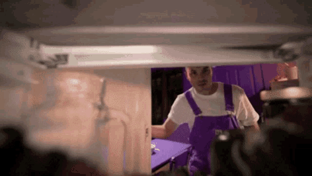 a man wearing purple overalls and a white shirt is standing in a room .