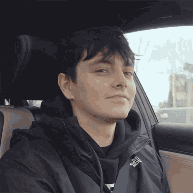 a young man wearing a black north face jacket is sitting in the driver 's seat of a car