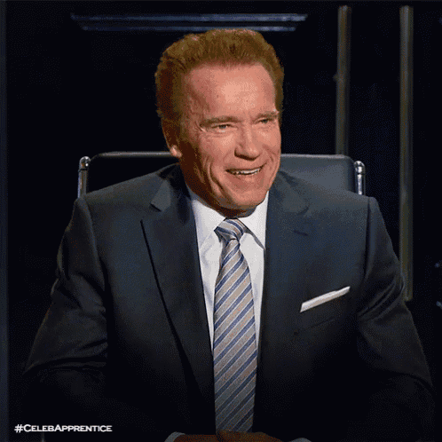 arnold schwarzenegger in a suit and tie smiles while sitting in a chair