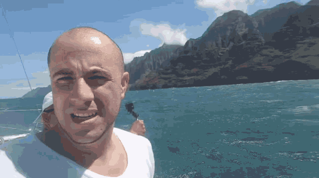 a man in a white shirt stands in front of a body of water with mountains in the background