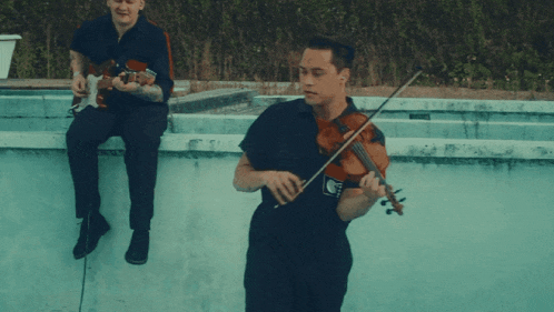 a man is playing a violin while another man sits on a wall playing a guitar