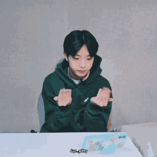 a boy in a green hoodie is sitting at a table with a cell phone and a pop socket