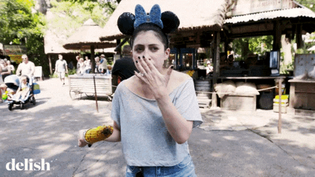 a woman wearing a mickey mouse ear covering her mouth while eating corn on the cob