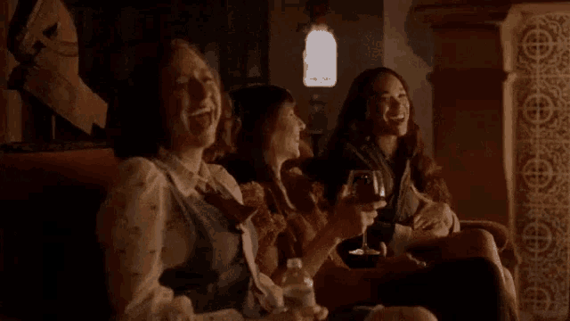 three women are sitting on a couch laughing and holding wine glasses .