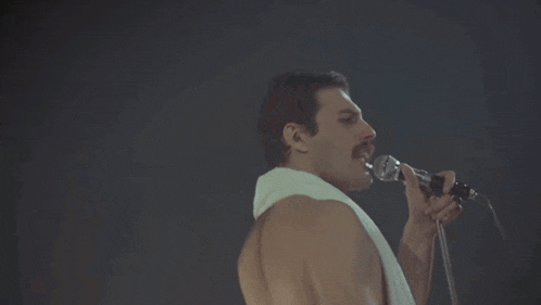 a shirtless man singing into a microphone with a white towel around his neck