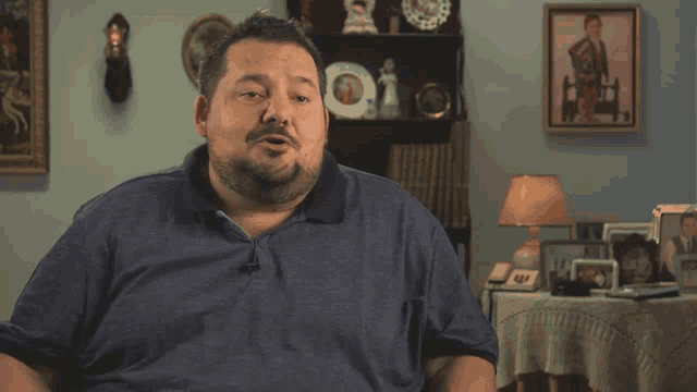 a man in a blue shirt is sitting in a living room with pictures on the wall and a lamp