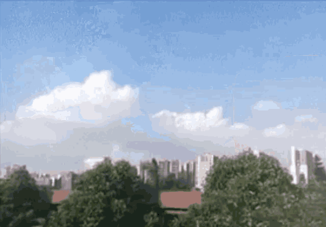 a city skyline with trees in the foreground and a blue sky with white clouds
