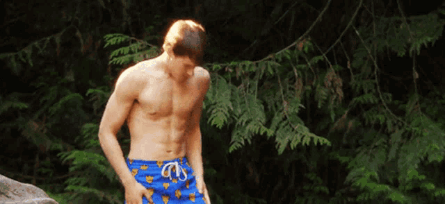 a shirtless man in blue swim trunks stands in front of trees