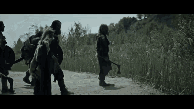 a group of men are walking down a dirt road holding axes and shields