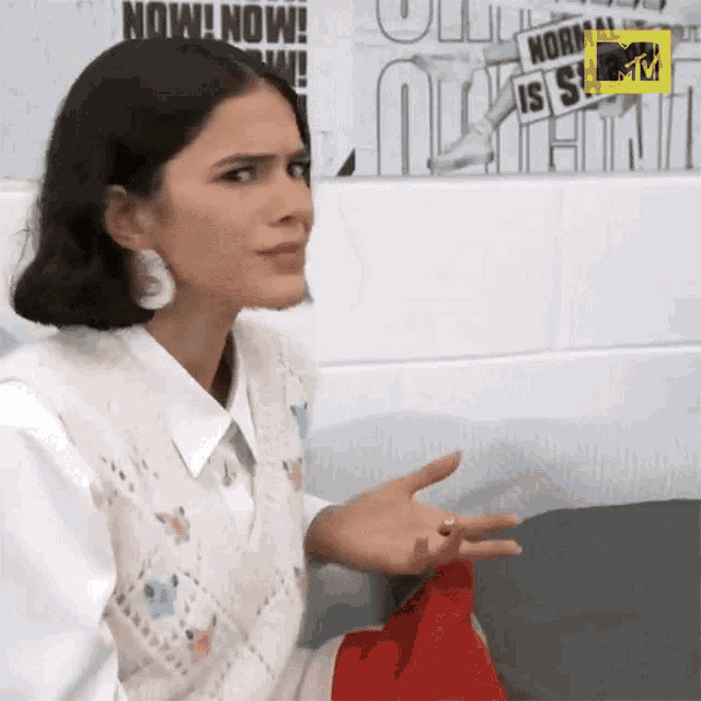 a woman wearing a white shirt and a white vest is sitting on a couch with a mtv logo on the wall behind her