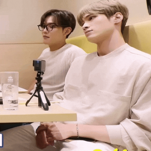 two young men sit at a table with a camera on a tripod that says ' nikon ' on it