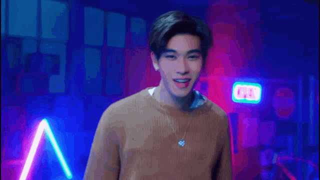 a young man wearing a brown sweater and a necklace is standing in front of neon lights .