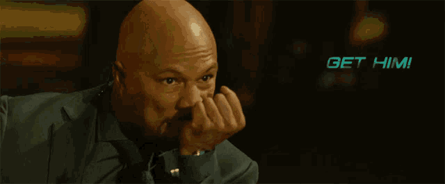 a bald man in a suit and tie is holding his fist to his mouth with the words " get him " behind him