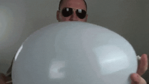 a man wearing sunglasses is blowing up a white balloon with a straw .