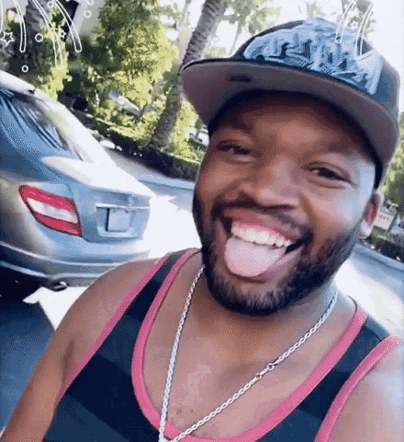 a man wearing a hat and a tank top is sticking his tongue out while standing in front of a car .