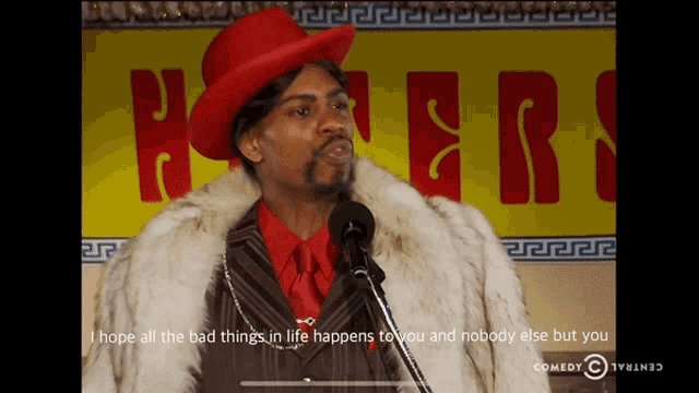 a man in a red hat and fur coat stands in front of a yellow sign that says haters