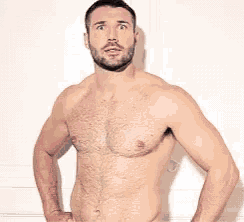 a shirtless man with a beard is smiling while standing in front of a wall .