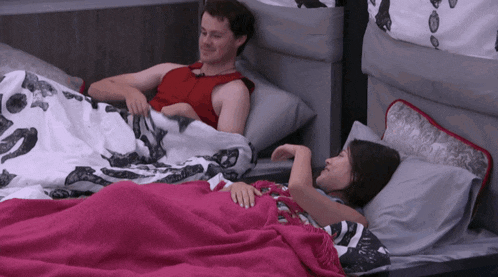 a man in a red tank top sits next to a woman in a bed under a pink blanket